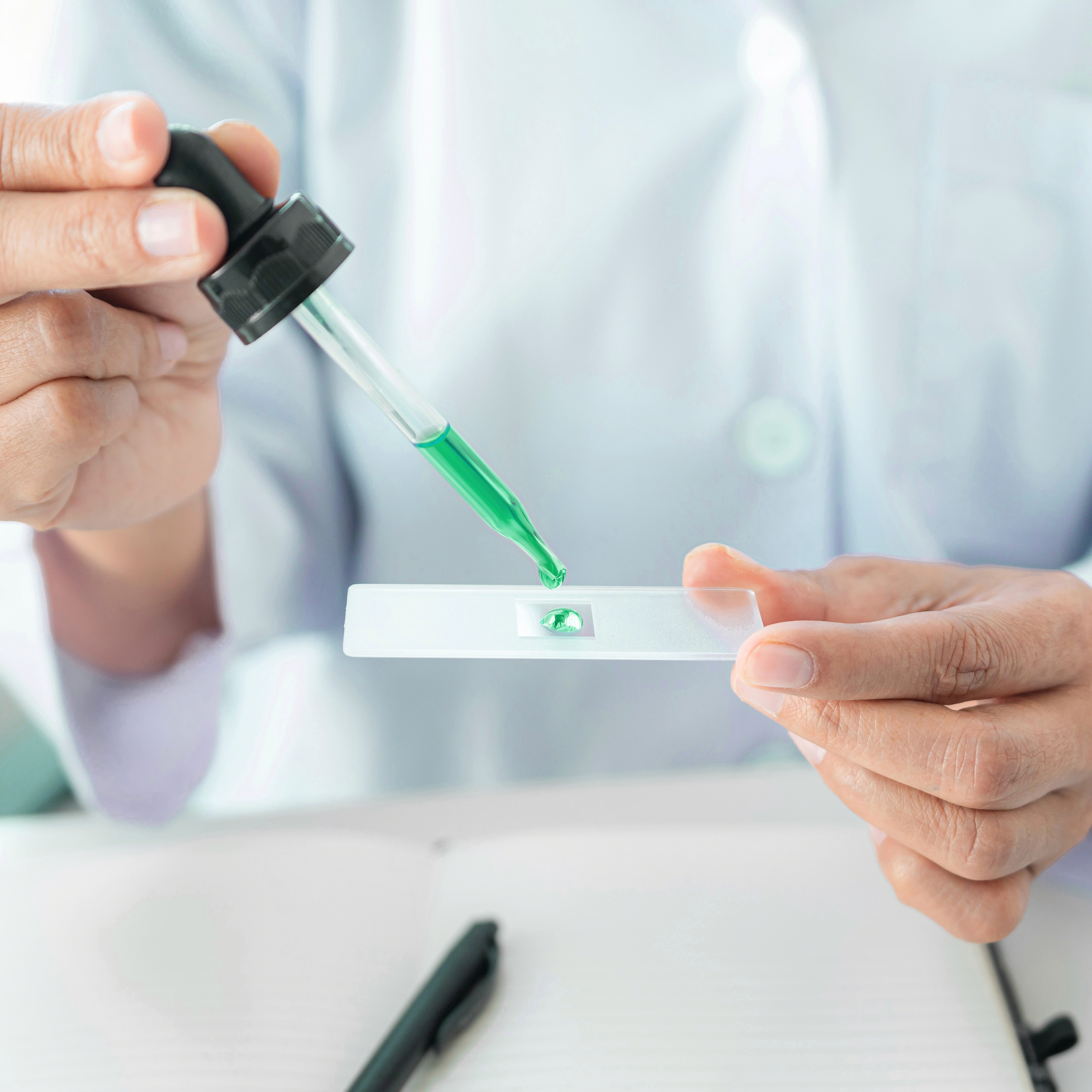 A person uses a dropper to place green liquid onto a glass slide, likely for a scientific or medical experiment.