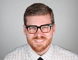A man with light brown/reddish hair and a reddish-brown beard is smiling at the camera. He wears black-framed glasses and a light-colored plaid shirt with a dark tie. The background is a solid light gray.
