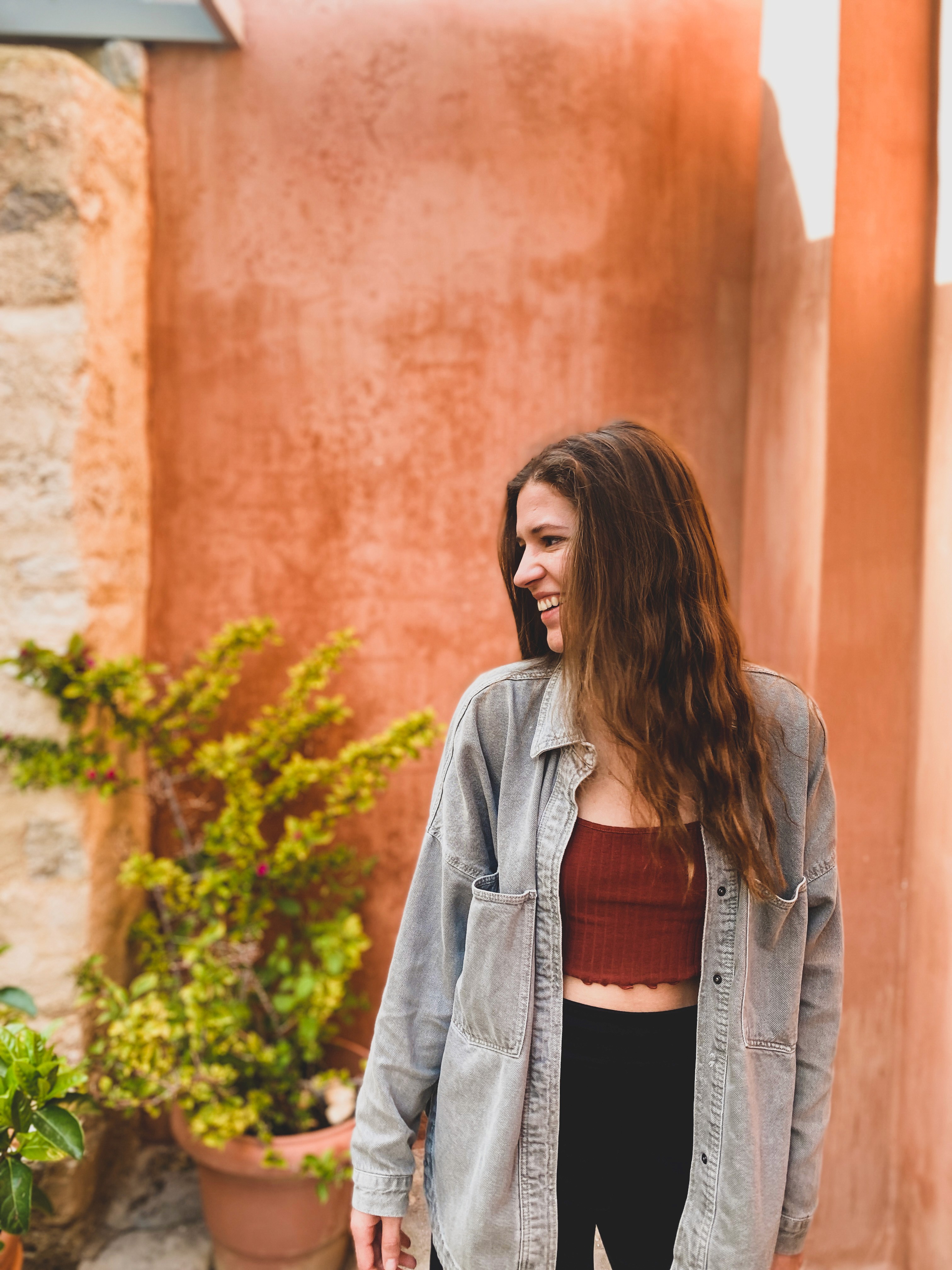 A young woman with long hair stands smiling near a potted plant against a colorful wall.