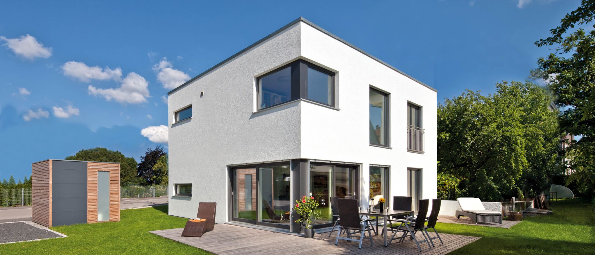 Modernes, zweistöckiges weisses Haus mit grossen Fenstern und Balkon, davor Holzterrasse mit Gartenmöbeln und Liegestuhl.