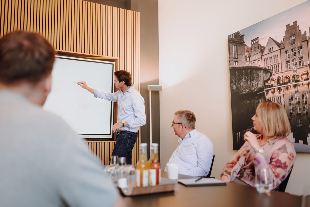 Ein männlicher Geschäftsführer präsentiert Inhalte an einem Whiteboard vor einer sitzenden Gruppe im Besprechungsraum von SLB.