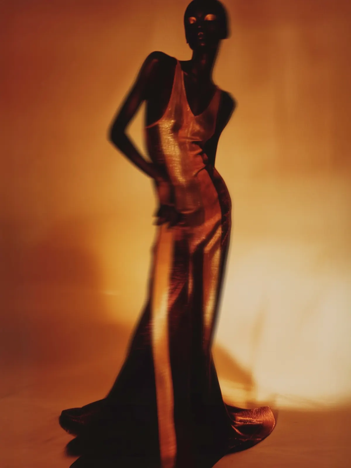 Full-length fashion portrait in a long gold dress against a warm studio backdrop.