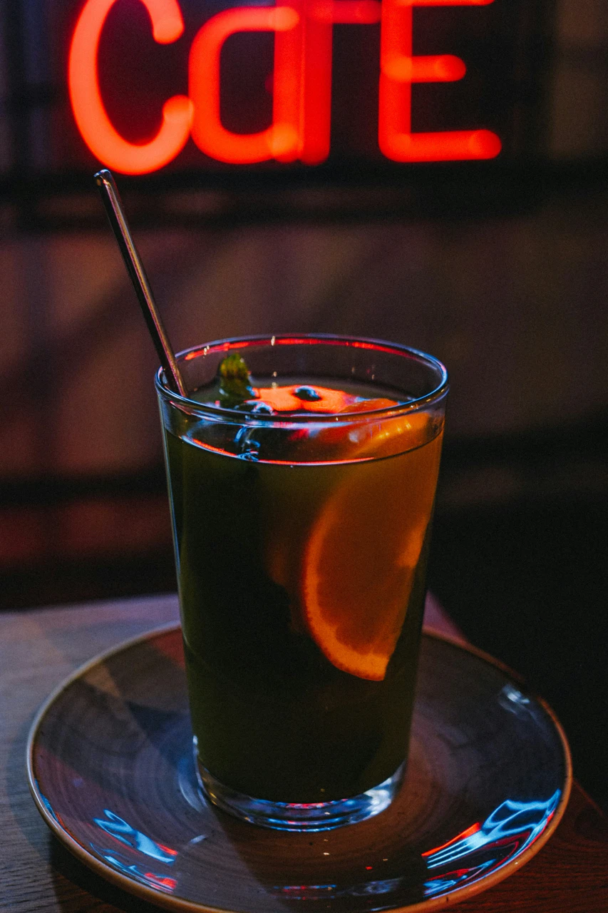 Glas mit einem Getränk, dekoriert mit einer Zitronenscheibe und einem Metallstrohhalm, vor unscharfem Hintergrund mit neonrotem Schriftzug.