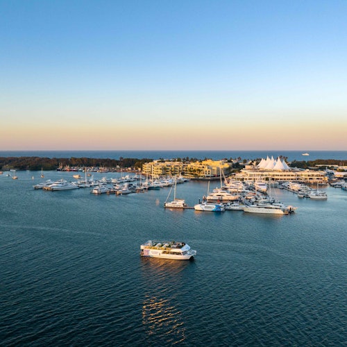 Aerial view of a marina with numerous boats docked, a ferry sailing in open water, and buildings on the shoreline at sunset.