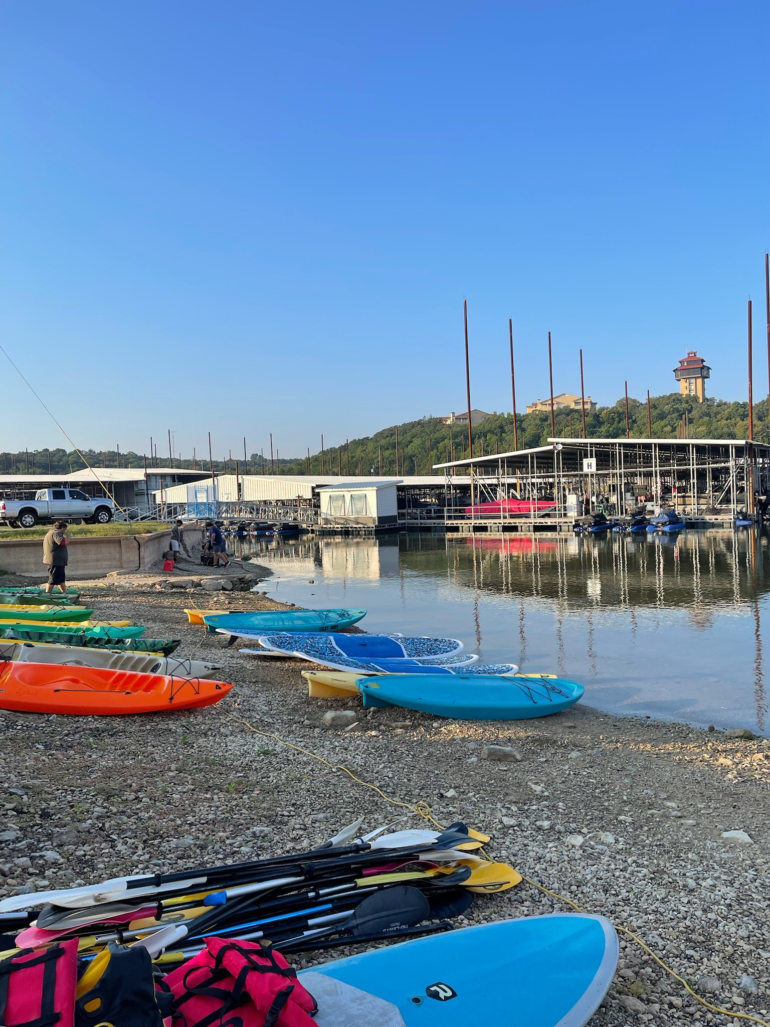 A serene lakeside scene features vibrant kayaks and paddleboards lining the pebbled shore, with calm water reflecting the clear blue sky and distant marina structures under a gentle morning light.