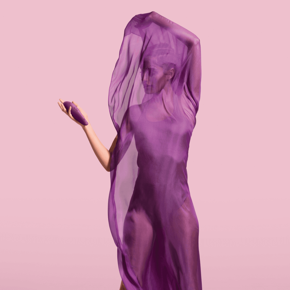 Womanizer Kampagnenmotiv – Frau in fliessendem violettem Chiffon-Schleier vor rosa Hintergrund, hält das Produkt in der ausgestreckten Hand.
