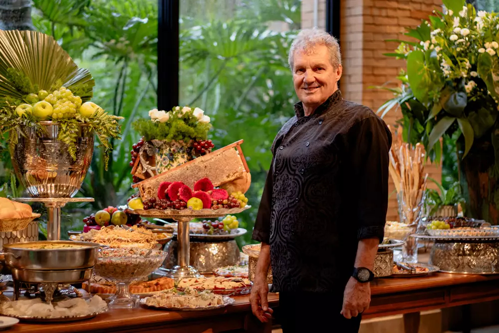 Chef Nenê Mangolini com o fundo decorado e diversos alimentos do buffet