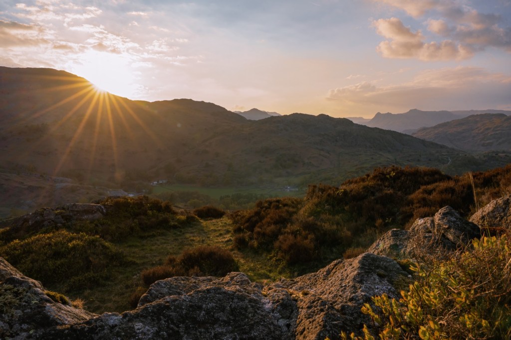 The setting sun creates a sun star behind the Wetherlam mountain range. Purple-pink heather is in the foreground.