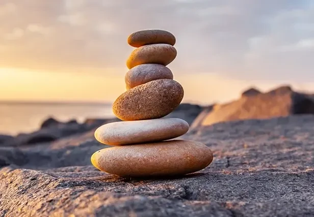 A stack of six smooth, rounded balancing stones sits on a dark, textured rock surface against a soft, golden sunset over the ocean.