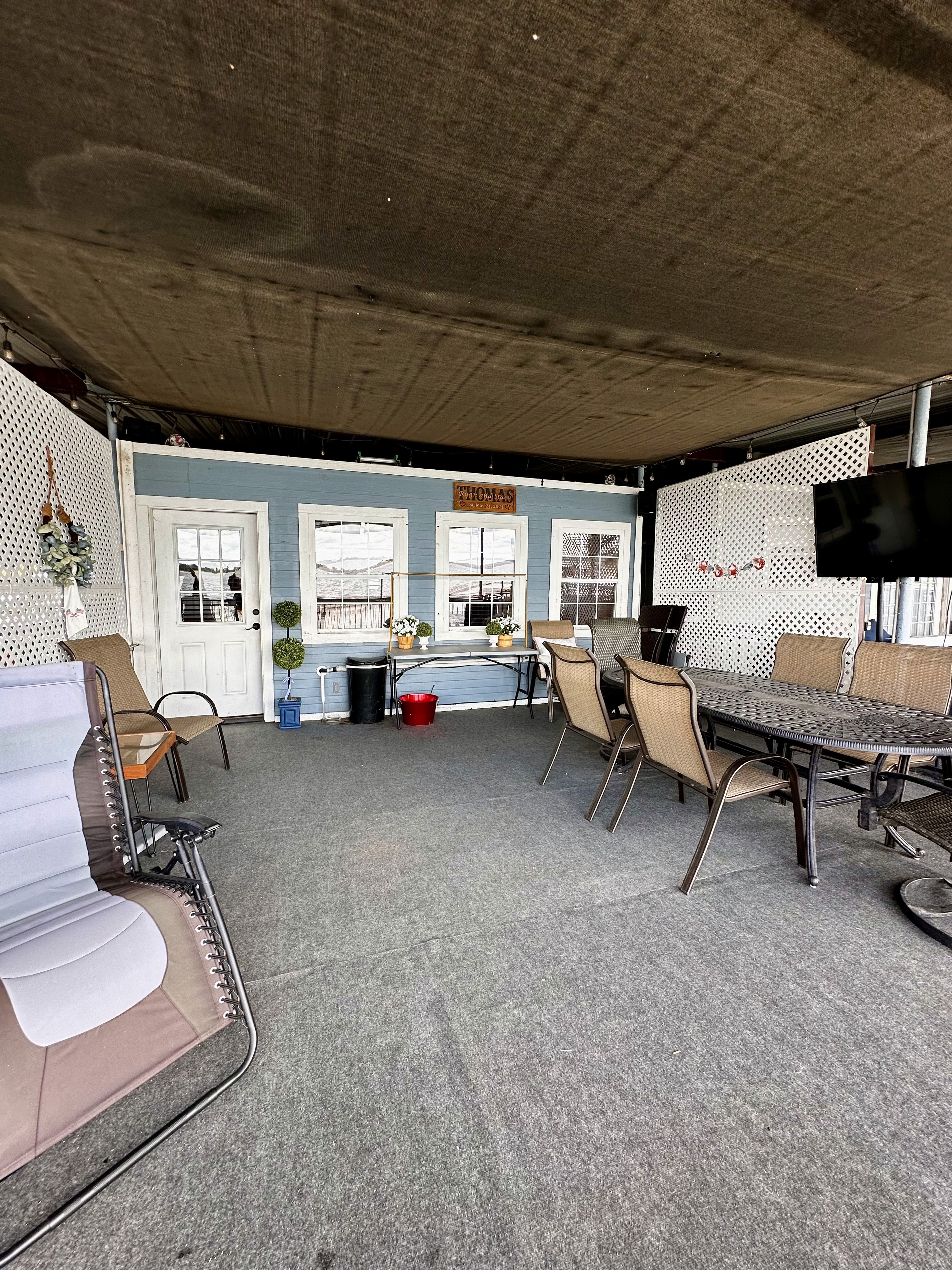 An outdoor covered patio features a cozy seating area with brown wicker chairs around a glass-top dining table, complemented by a white lattice backdrop and shaded by a brown fabric canopy.