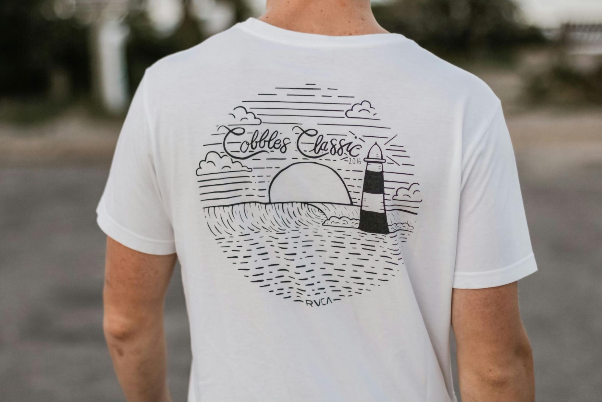 The back of a person wearing a white t-shirt. The t-shirt features a black design that depicts a lighthouse, a setting sun, and some clouds. The words 'Colblos Classic' are written in cursive script at the top of the design. The person is standing outdoors, with a blurred background that suggests a natural setting, possibly a park or beach.