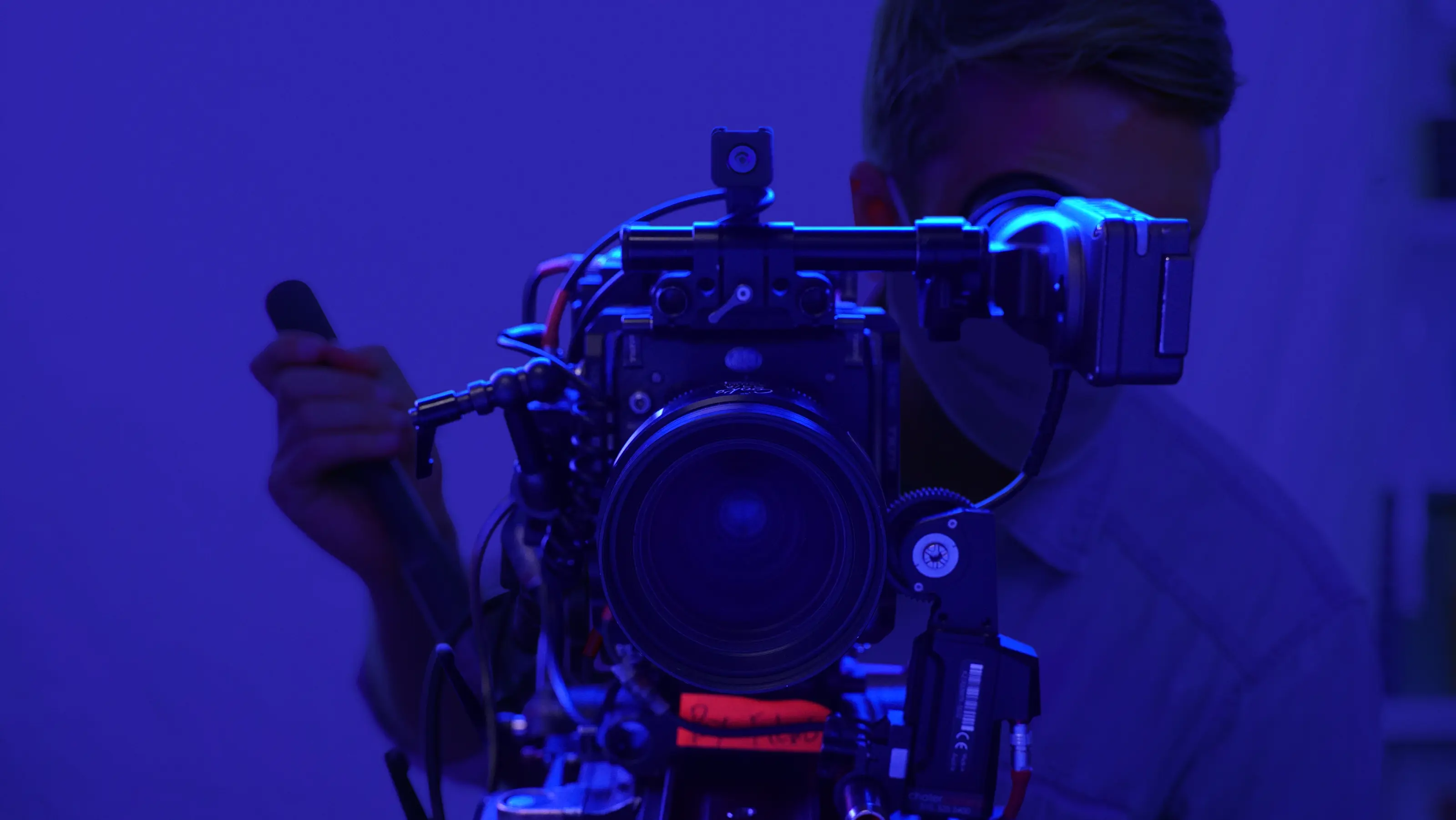 A person holding a camera in a dimly lit setting with a blue hue, focusing on a subject off-screen.