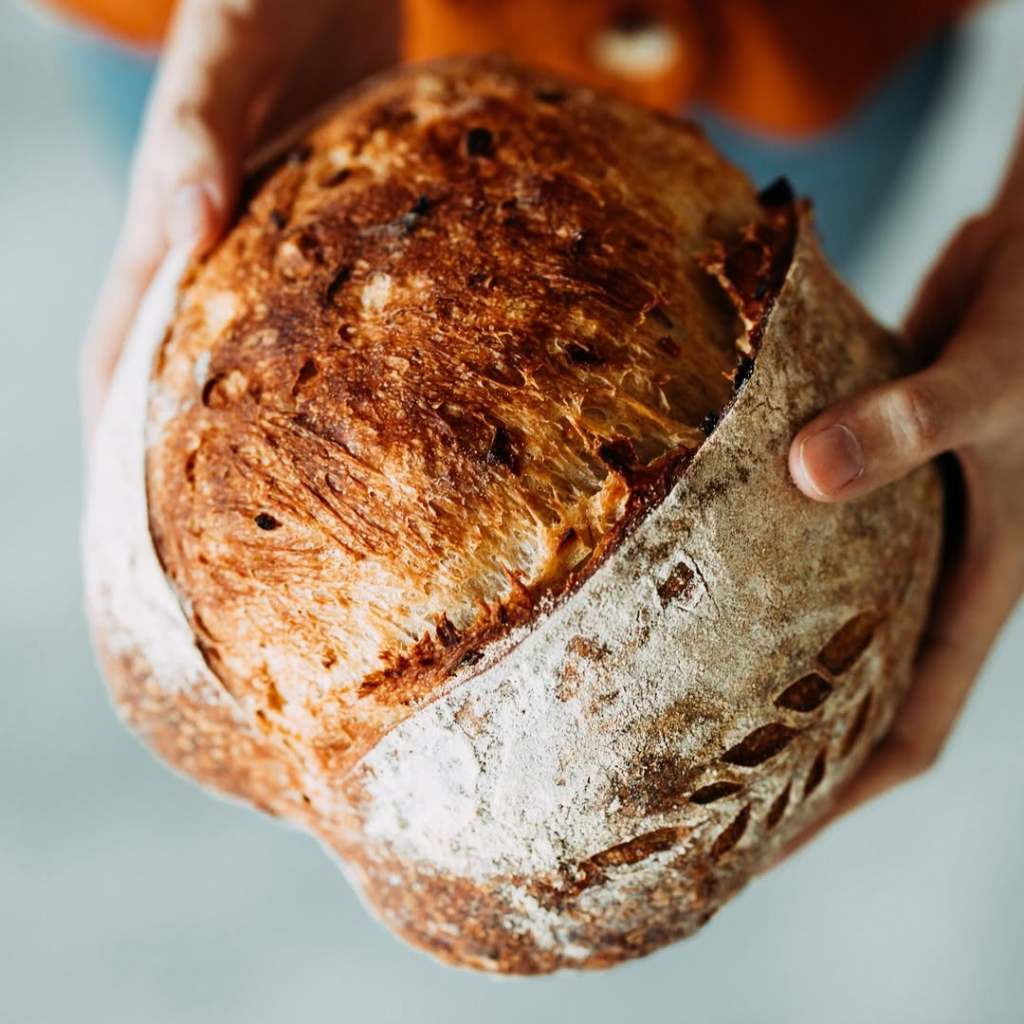 A person holding a freshly baked sourdough bread with a golden-brown crust and a rustic design.