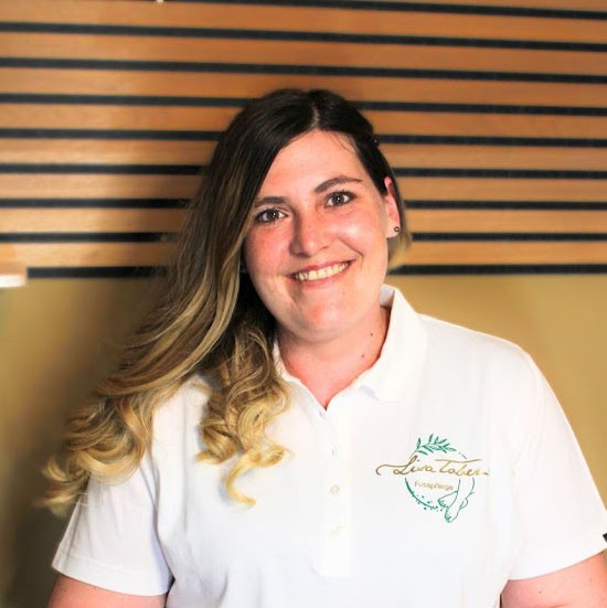 A smiling woman with her arms crossed, standing against a dark green background. She has long, dark hair.