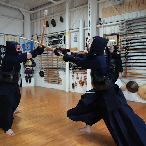Twee personen in traditionele kendo-kleding sparren met bamboezwaarden in een dojo, waar verschillende martial arts uitrustingen aan de muren hangen.