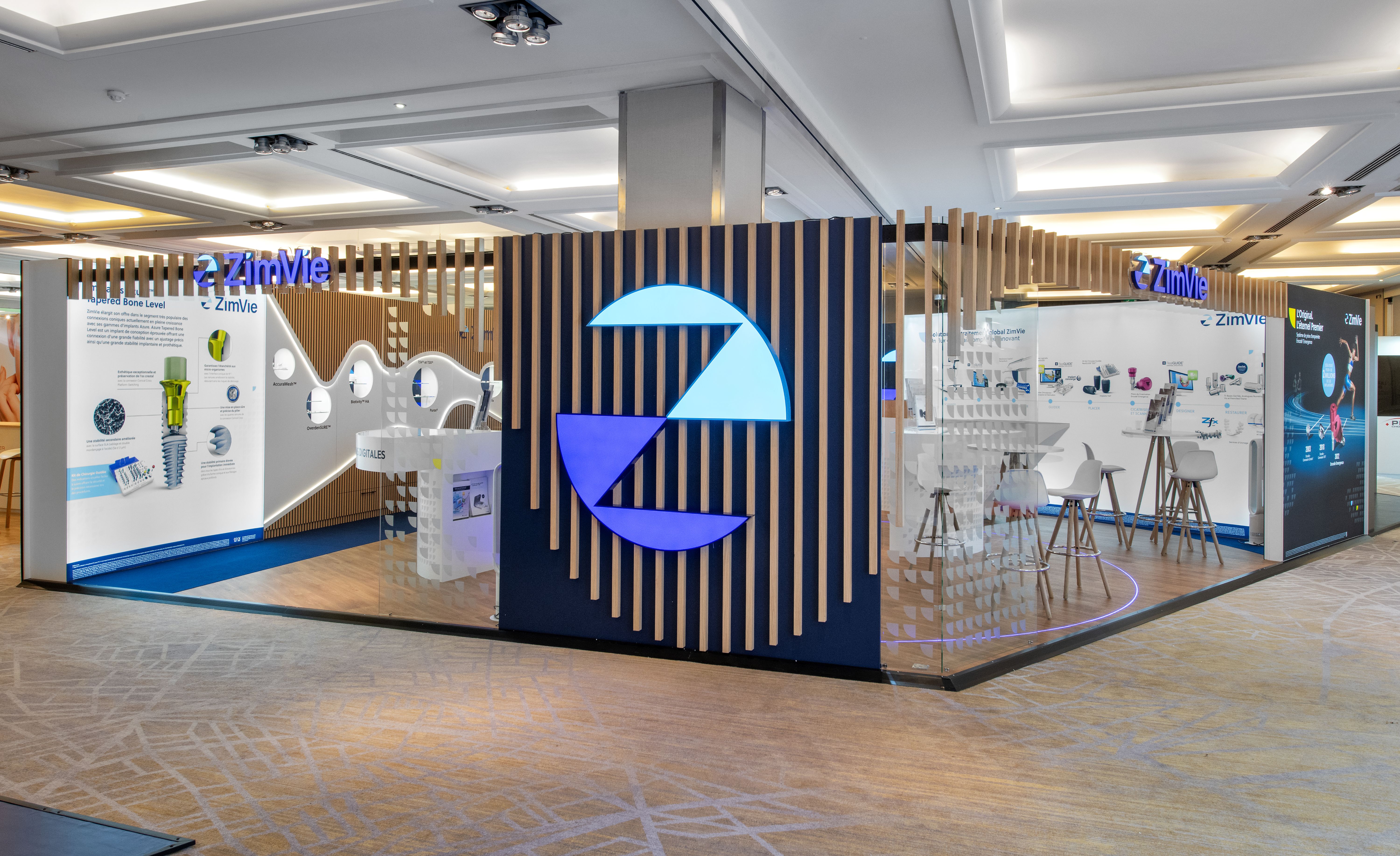 Stand ZimVie ADF 2025 par Expace – vue façade avec colonne centrale logo rétroéclairé bleu marine, lames bois verticales, zones Solutions Digitales et Biomatériaux et panneaux produits implants dentaires