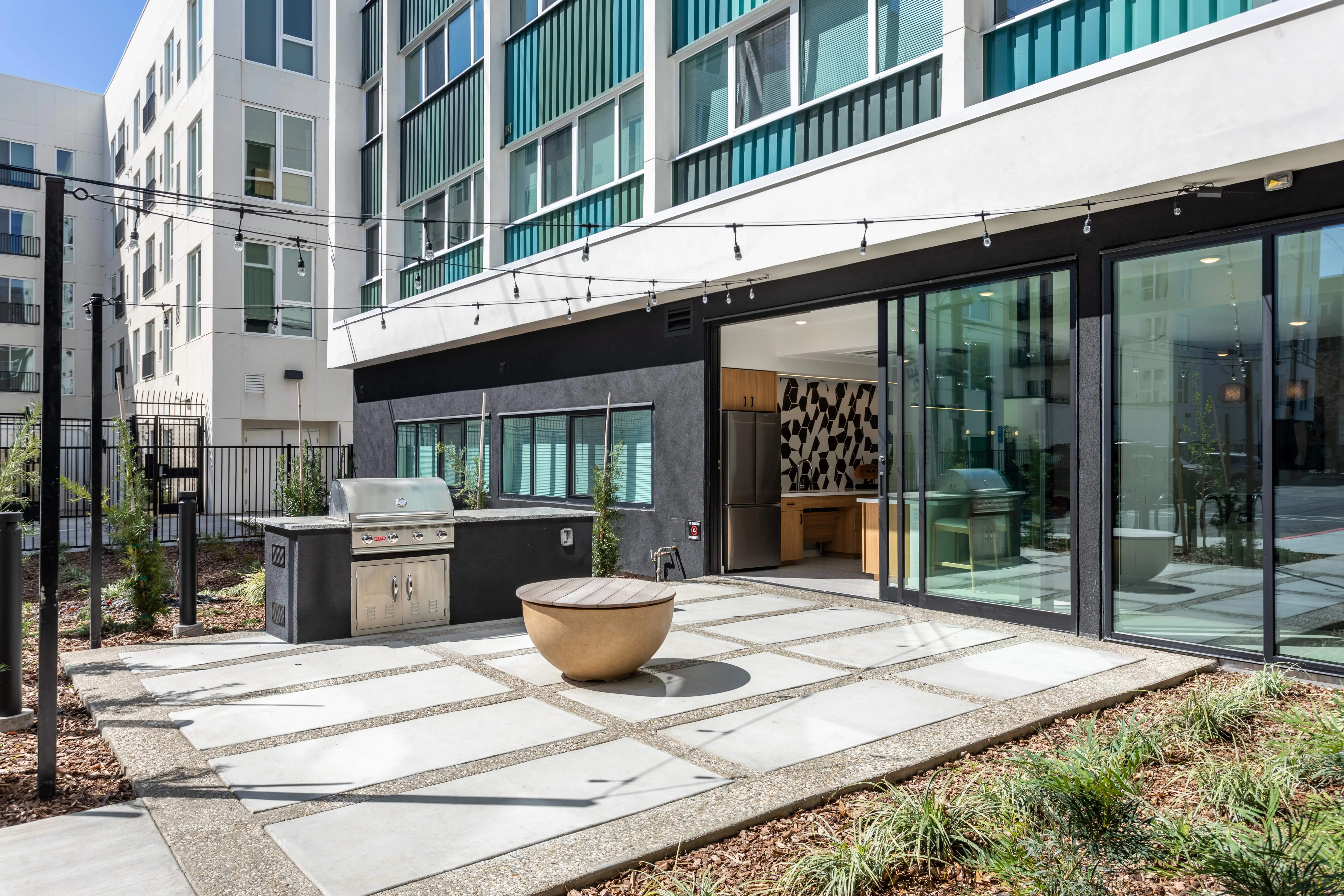 Outdoor communal area of a modern apartment complex in Midtown Sacramento featuring a sun-drenched barbecue grill, string lights, a round fire pit, and a path with large concrete slabs. Large glass doors provide access to an indoor space with contemporary decor and modern living spaces.