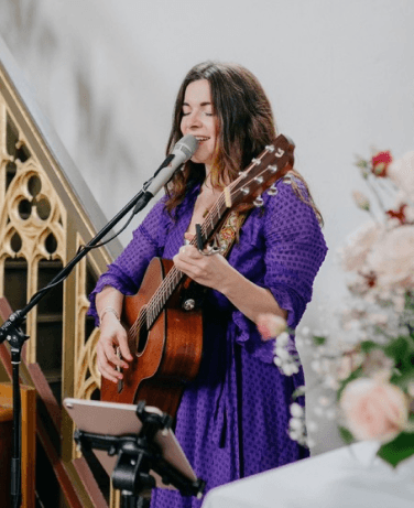 Maria Wiethaler freie Rednerin und Sängerin für Hochzeiten, Taufen, Trauerfeiern und Events aus München und Landshuter Umgebung. Eine Sängerin in einem lila Kleid steht vor einem Mikrofon und spielt eine Akustikgitarre. Sie wirkt vertieft in ihre Musik, mit geschlossenen Augen und einem sanften Lächeln. Im Hintergrund sind florale Dekorationen und gotische Architekturelemente sichtbar, die eine festliche und elegante Atmosphäre vermitteln.