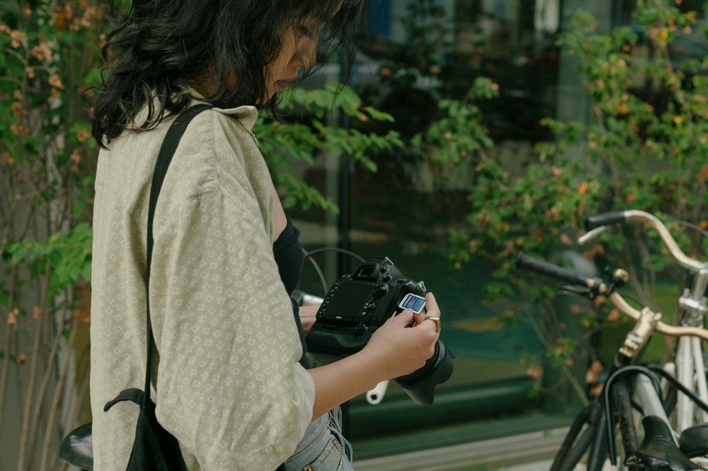 A young woman stands outdoors, carefully adjusting her digital camera, ready to capture photographs, with a backdrop of greenery and bicycles, highlighting her focus and passion for photography.