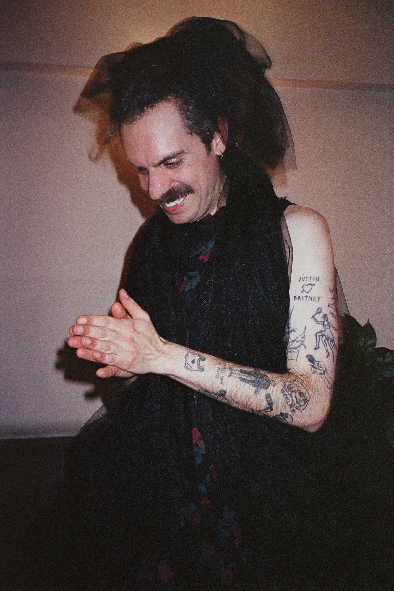 A person with tattoos and a mustache is smiling while wearing a black veil and sleeveless outfit, clasping their hands together in front of a neutral background.