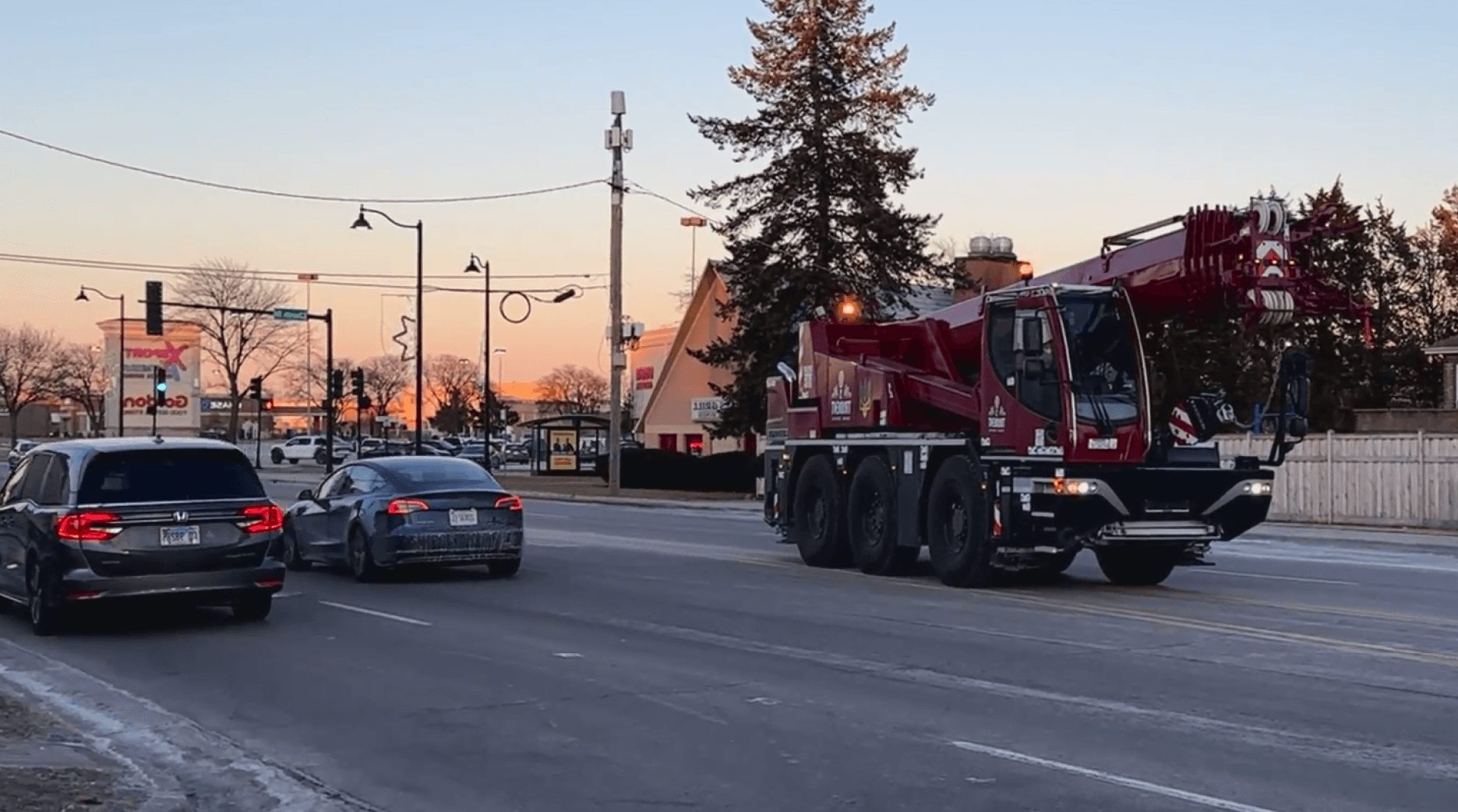 A large red crane truck travels on a suburban road at dusk, passing by two cars on the left, with a backdrop of bare trees, streetlights, and a fast-food restaurant, under a clear evening sky.