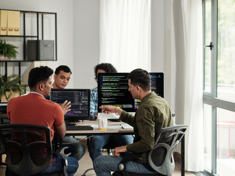 Scoped Work - Three people sit around a table discussing work, with laptops and monitors in a bright, modern office space.