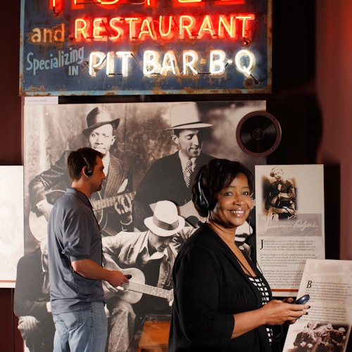 Dva lidé na výstavě, kde je vystavený neonový nápis a historická fotografie hudebníků. Žena se usmívá, drží zařízení, zatímco muž sleduje expozici.