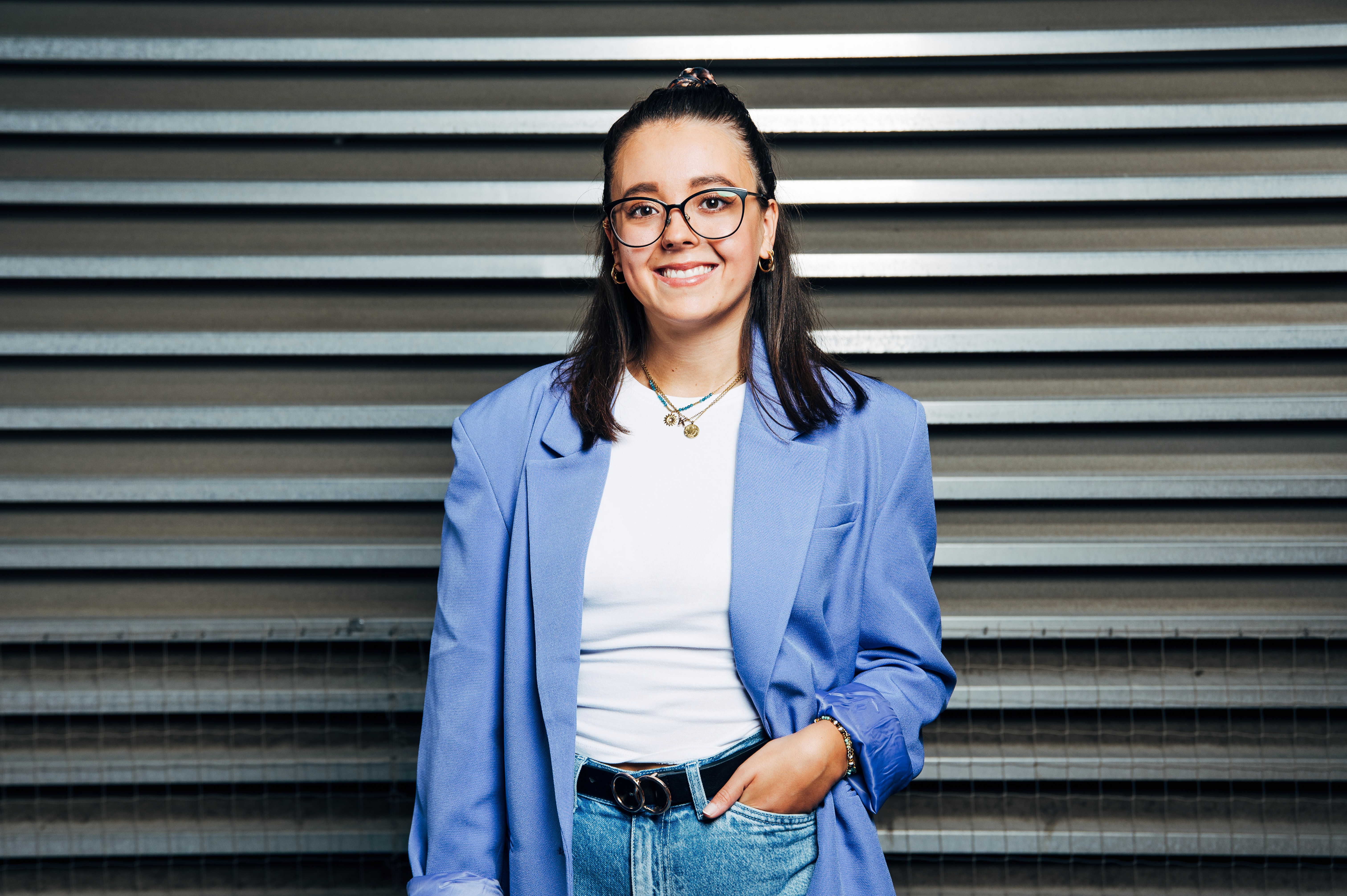 Smiling person wearing glasses and a blue blazer, standing against a striped wall.