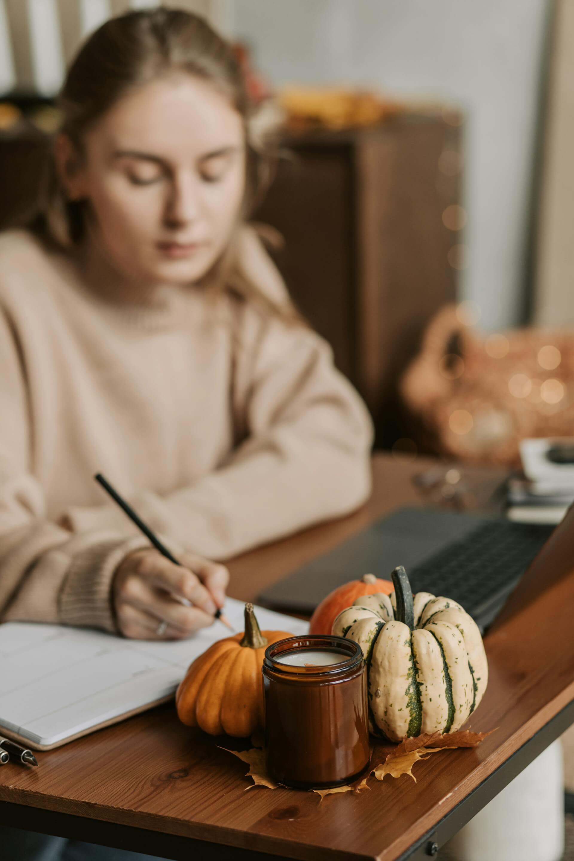 A woman writes in a notebook at a table, accompanied by a laptop and decorative pumpkins