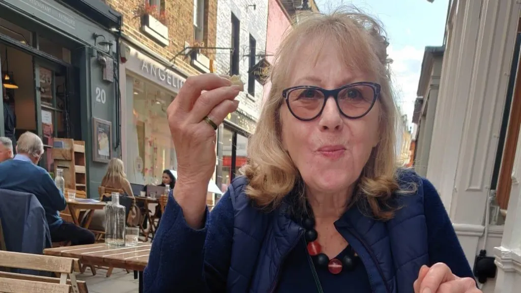 Susan Copping smiling while eating at an outdoor café, enjoying a relaxed moment on a sunny street.