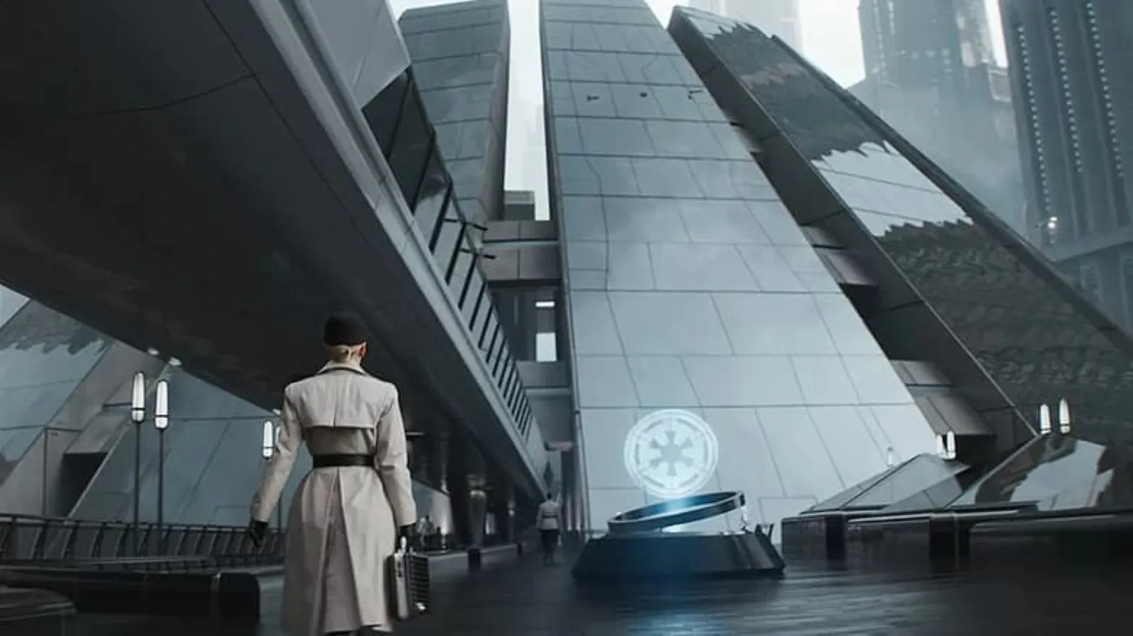 A person in a long, white coat walks under an enclosed futuristic bridge as they approach a futuristic, metallic building with angular architecture and an emblem on a circular platform, set against a foggy cityscape.