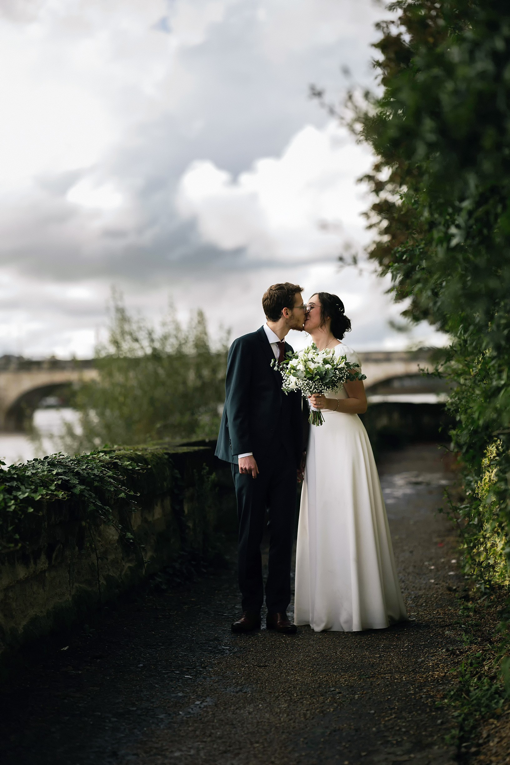 A couple in wedding attire shares a romantic kiss on a leafy path by a tranquil river.
