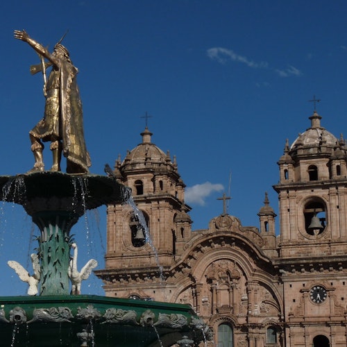 一座銅製雕像矗立在水泉上，位於一座歷史悠久的教堂前，教堂有兩座鐘樓，在晴朗的藍天下。