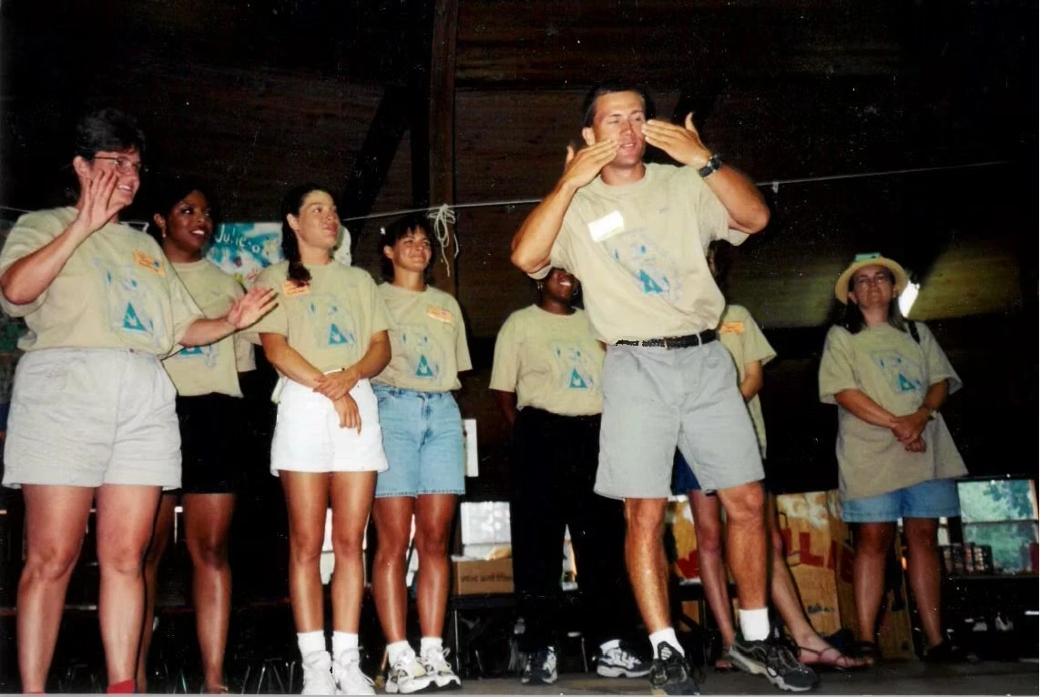 A group of camp staff stand on a stage wearing matching tan shirts. One man in the center is signing to a crowd.