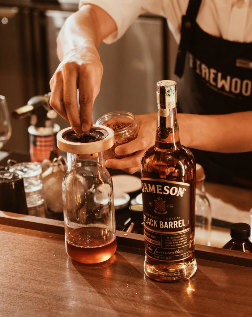 A bartender adding spices to a decanter containing Jameson Black Barrel whiskey, with the bottle prominently displayed on the bar.