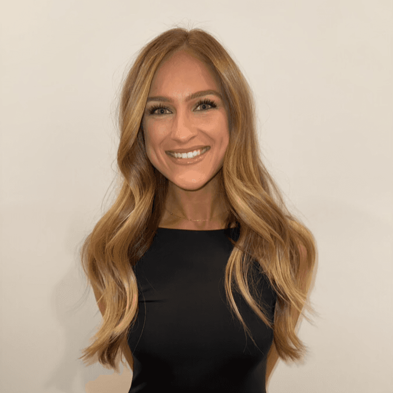 A woman with red hair wears a lavender coat and a white sweater, accessorized with a gray scarf. She has a calm expression against a neutral background.