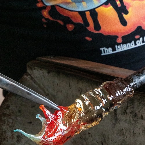 一张玻璃吹制过程的特写,炽热的熔融玻璃在缤纷的背景前正借助工具进行塑形。