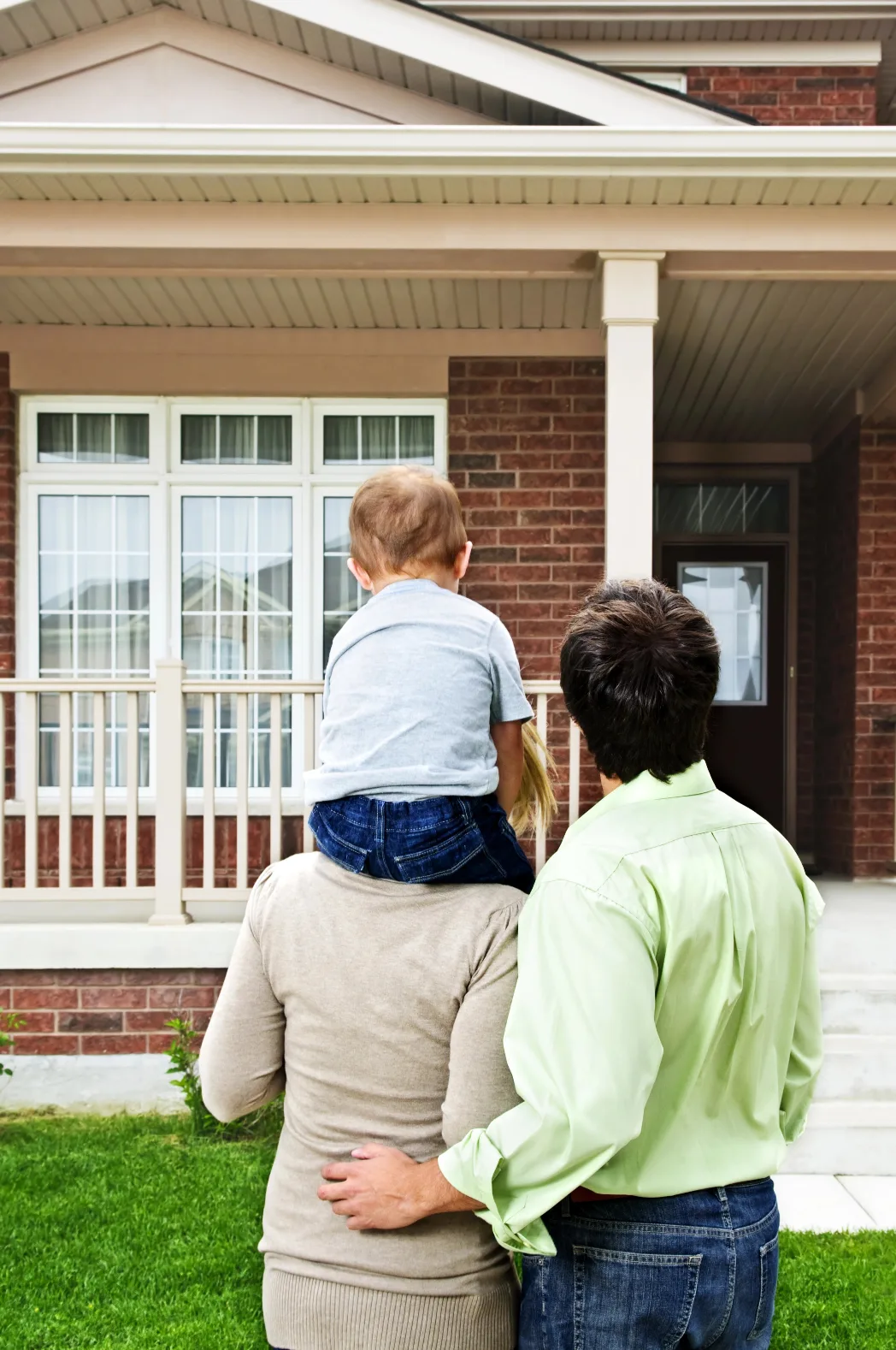 Two adults and a toddler view the front exterior of a brick home with white-trimmed windows and a covered porch.