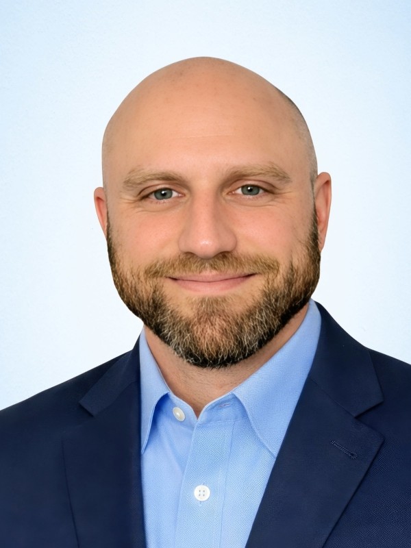 A man in a black suit and tie stands confidently with arms crossed against a gray background. He projects professionalism and determination.