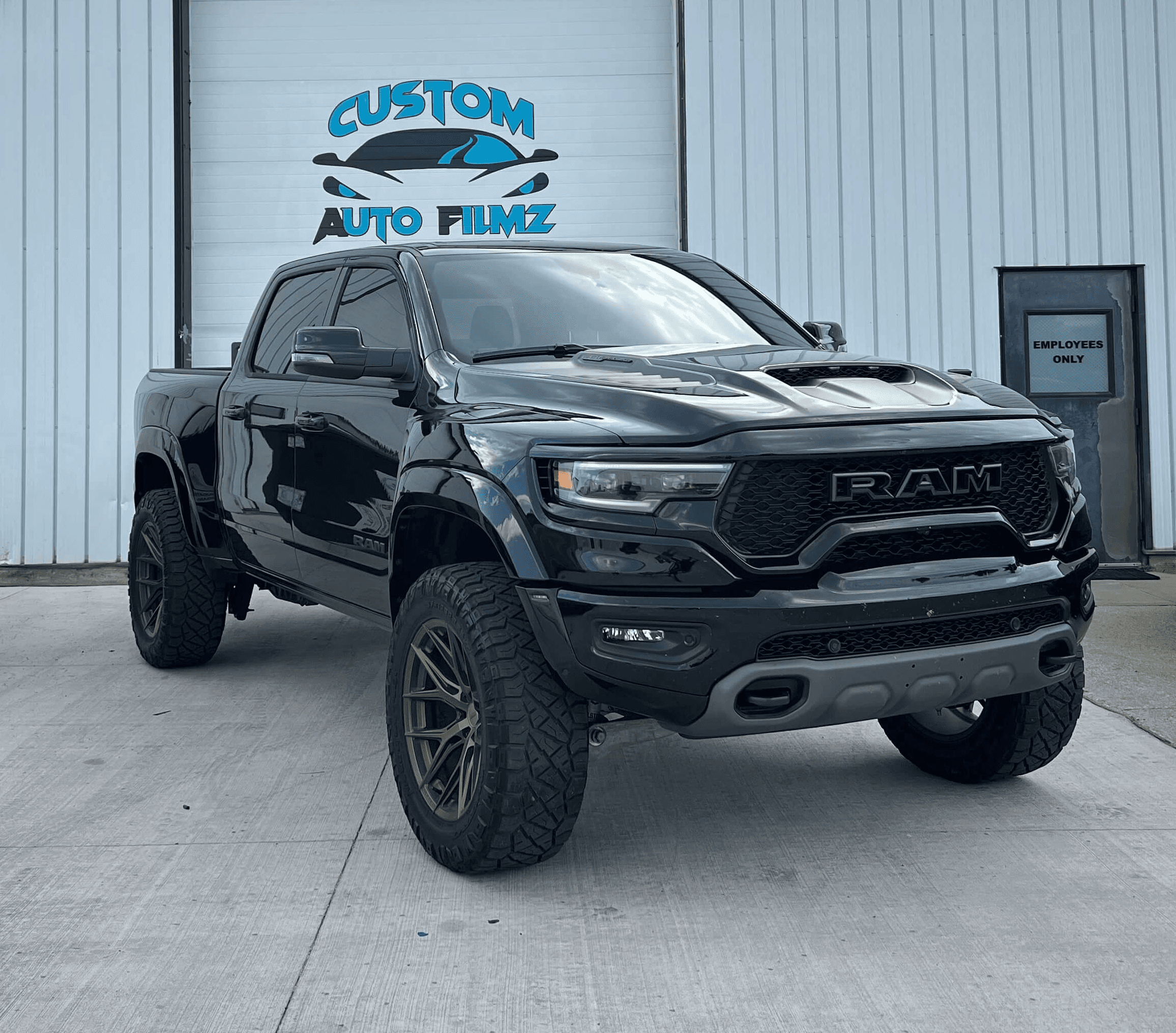 A black RAM 1500 TRX pickup truck is parked on a concrete driveway in front of a white industrial building.