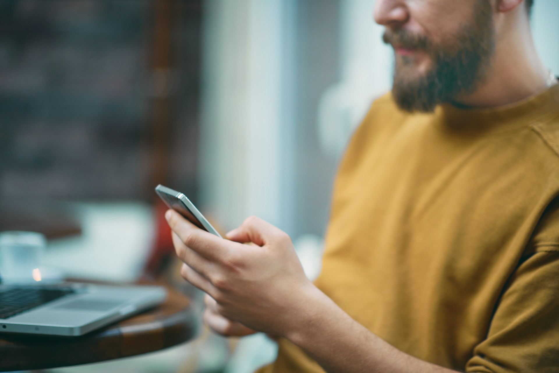 A man sitting indoors, holding a smartphone in one hand with a laptop on the table beside him, partially out of focus.