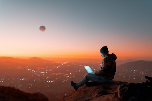 A person sitting on a ledge, working on a laptop against a vibrant sunset backdrop.