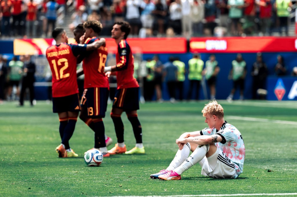 A dejected Belgium player sits on the grass while Spanish teammates celebrate in the background at the 2026 World Cup