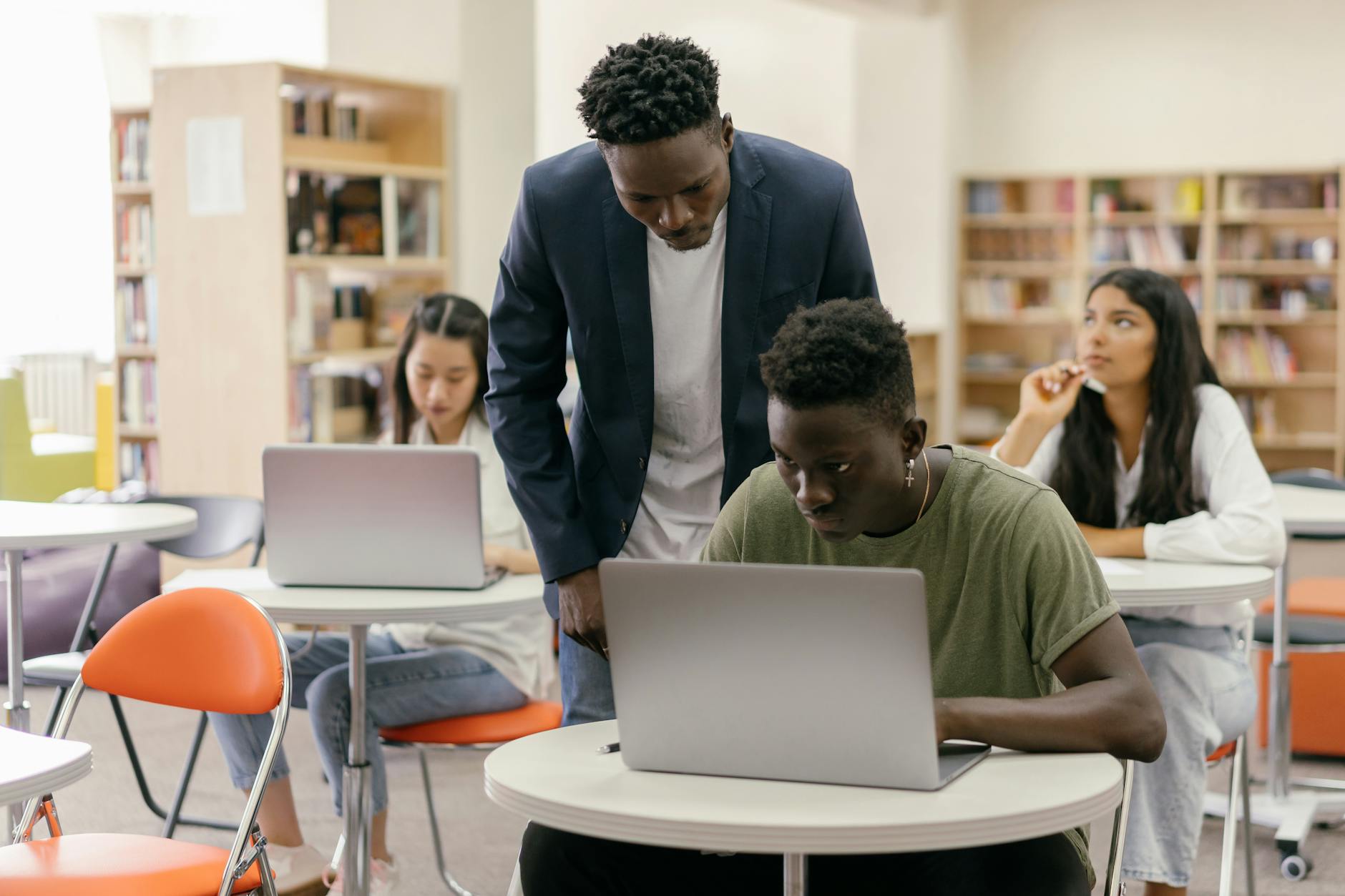A frustrated teacher looking at a laptop screen while students in the background are distracted by their smartphones.