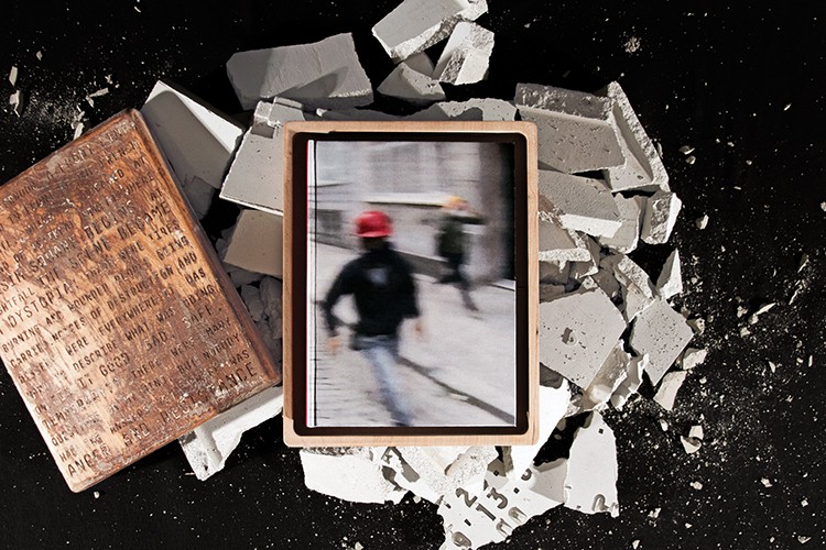 The image shows a rectangular frame with a light brown or beige border, containing a blurred photograph. The photo shows two people walking on a light-colored paved surface; one person wears a red hard hat. The surrounding area is filled with broken pieces of concrete and stone in various sizes and colors, scattered on a dark surface. A portion of a book with a worn and faded cover is also visible in the bottom left of the image.