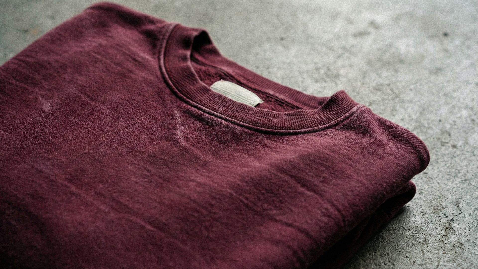 A close-up macro detail shot of a garment-dyed heavyweight fleece hoodie in rich, uneven vintage-washed burgundy on a raw concrete surface, showing the depth of the dye and the quality of the fleece with a small woven label partially visible at the collar.