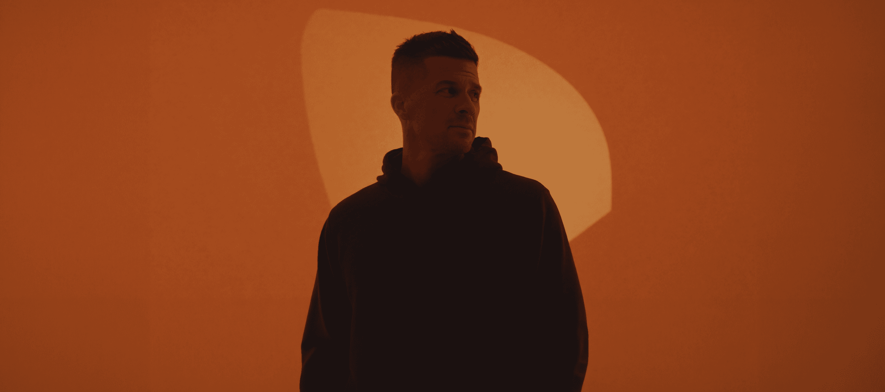 A person stands against a vibrant background of orange and yellow wavy patterns. They are wearing a dark coat, looking contemplative and thoughtful.