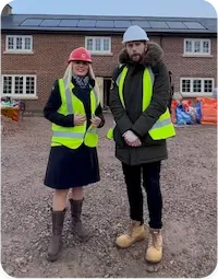 Two people wearing high-visibility vests and hard hats standing on a construction site in front of a new brick house development