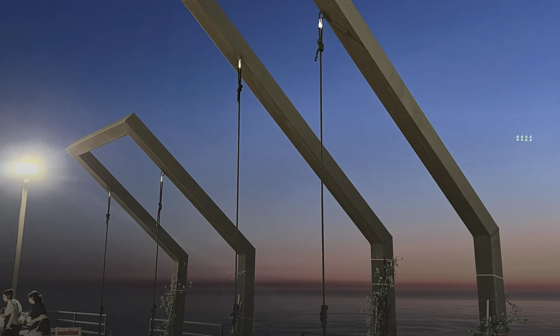 Modern architectural workspace with large floor-to-ceiling windows, surrounded by trees, and desks filled with design materials and books. Two blurred figures are walking through the space.