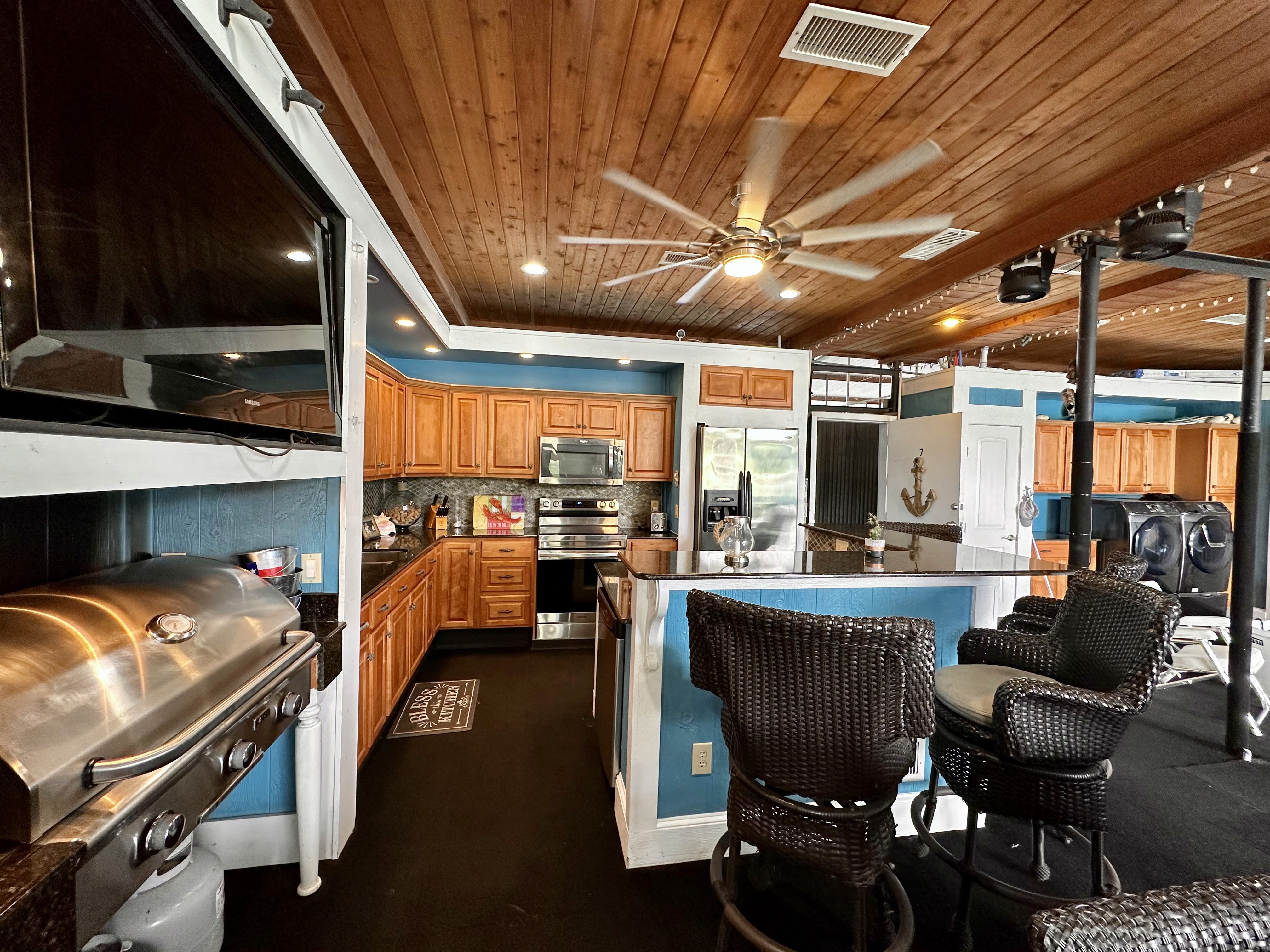 A cozy kitchen features a polished wooden ceiling, navy blue walls, and a large island with wicker bar stools, complemented by stainless steel appliances and decorative nautical accents.